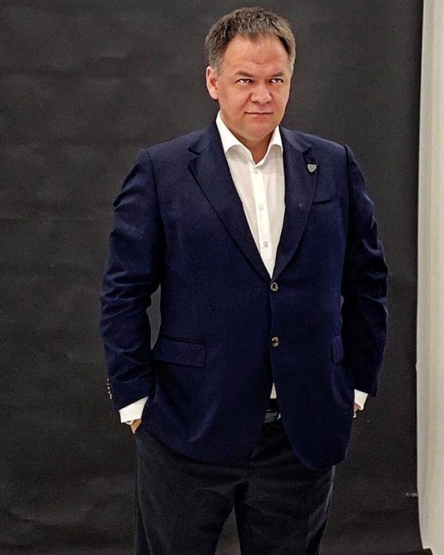 Александр Копытько в пиджаке, руки в карманах, нейтральный фон студии